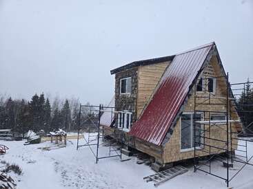 A snowy scene shows an A-frame house under construction. Scaffolding surrounds the building. Trees line the background, as snowfall gently covers the ground and roof.