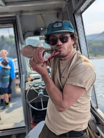 A man on a boat playfully kisses a freshly caught fish, wearing sunglasses and a blue hat. Water and other people are visible in the background.