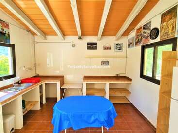 The image depicts a small kitchen with white walls, wooden ceiling, and brown tiled floor, featuring a sink, table, and refrigerator.