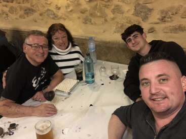 The image shows a group of four people sitting at a table with drinks and food, posing for a selfie in a rustic setting.