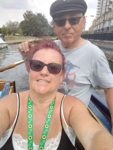 The image shows a selfie of a man and woman on a boat, with the woman in the foreground and the man behind her, both wearing sunglasses.