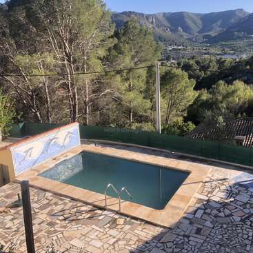 The image depicts a serene pool scene with a mountainous backdrop, featuring a rectangular pool, mosaic tiles, and a scenic view of trees and mountains in the distance.