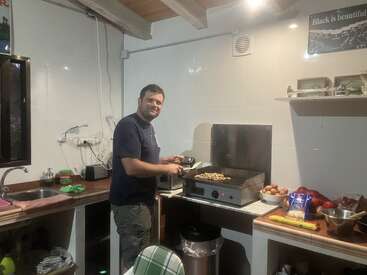 The image depicts a man cooking in a kitchen, standing beside a grill with food on it, wearing a dark blue t-shirt and green pants.