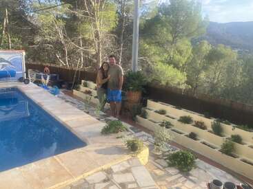 The image depicts a serene outdoor setting, featuring a pool, a couple posing for a photo, and a lush garden with trees and a mountainous backdrop under a clear blue sky.