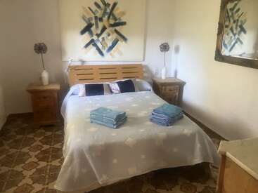 A cozy bedroom with a double bed, neatly folded blue towels, abstract wall art, wooden furniture, decorative vases, patterned floor tiles, and a large wall mirror.