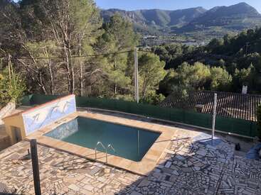 The image depicts a serene pool scene with a mountainous backdrop, featuring a rectangular pool, mosaic tiles, and a scenic view of trees and mountains in the distance.