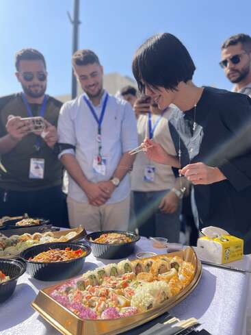 A group of people gathers outdoors, smiling and watching a woman using chopsticks to eat sushi from a beautifully arranged platter, enjoying a bright, sunny day.