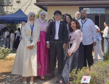 A group of people stand smiling outdoors, some dressed in traditional Korean and Middle Eastern attire, possibly celebrating a cultural event or international day at an institution.