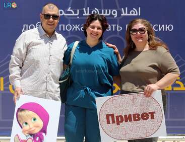 Three smiling people pose together outdoors, holding signs. One sign features a cartoon girl, the other says "Привет" (Hello in Russian). A blue welcome banner is behind them.