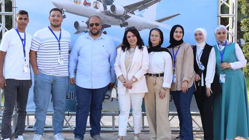 A group of eight people stand smiling in front of a backdrop featuring an airplane. They wear badges and casual-professional clothing, suggesting participation in an aviation event.