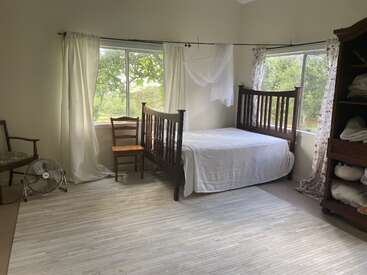 This cozy bedroom features simple wooden furniture, a white covered bed, two windows with curtains, a fan, an open shelf with linens, and soft natural light.