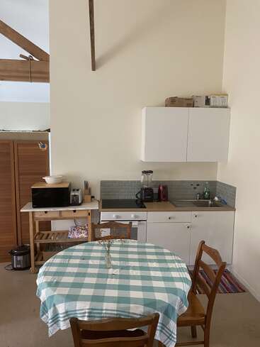 This image shows a small kitchen with white cabinets, a checkered tablecloth on a round table, wooden chairs, kitchen appliances, and simple modern decor.