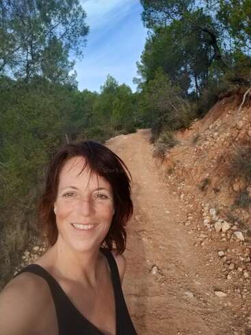 The image depicts a woman taking a selfie on a dirt path surrounded by trees and bushes, with a blue sky above.