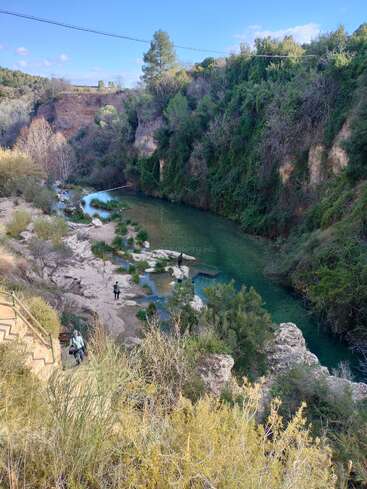 The image depicts a serene river flowing through a lush, rocky canyon, with people standing on the riverbank and a bridge spanning the waterway.