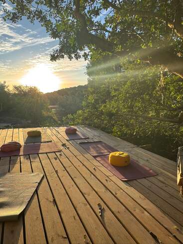 Sunrise illuminates a peaceful wooden deck set for yoga, with mats and meditation cushions beneath leafy trees, offering a serene natural setting for relaxation and mindfulness.