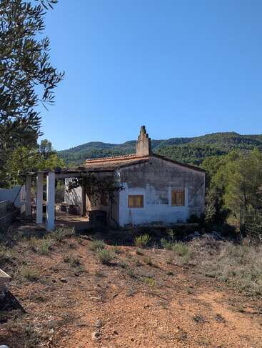 A small, rustic house with a red-tiled roof sits surrounded by dry, rocky ground and greenery, backed by forested hills under a clear blue sky.