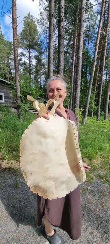 Una mujer sonriente está de pie al aire libre en un bosque, sosteniendo una cesta hecha a mano con asas de madera. Lleva un vestido marrón y zuecos negros, rodeada de árboles altos.