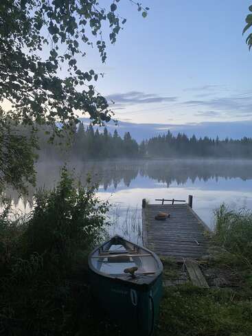 Una tranquila escena lacustre al amanecer, con niebla sobre el agua, un muelle de madera que se adentra en ella y una canoa descansando entre la exuberante vegetación de la orilla.