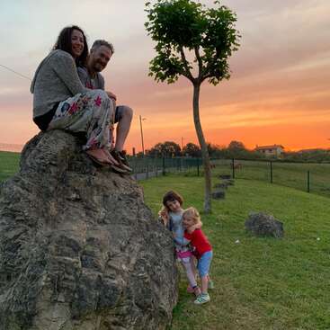 A imagem mostra uma família de quatro pessoas posando em uma grande rocha, com duas crianças abraçadas em sua base, em um cenário sereno de pôr do sol em um campo gramado.