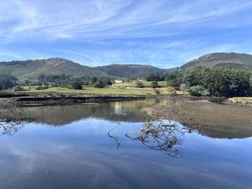 Um rio calmo reflete o céu azul, cercado por campos verdes, colinas e montanhas arborizadas. Um galho de árvore caído está parcialmente submerso na água.
