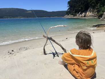 Uma criança com uma toalha laranja senta-se na areia da praia, observando uma vara de pescar apoiada em varas. O mar está calmo, com montanhas e árvores ao fundo.