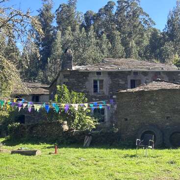 Uma antiga casa de pedra fica sob a luz do sol, cercada por uma vegetação exuberante. Uma faixa triangular colorida está pendurada sobre o quintal gramado, com uma cadeira e pneus por perto.