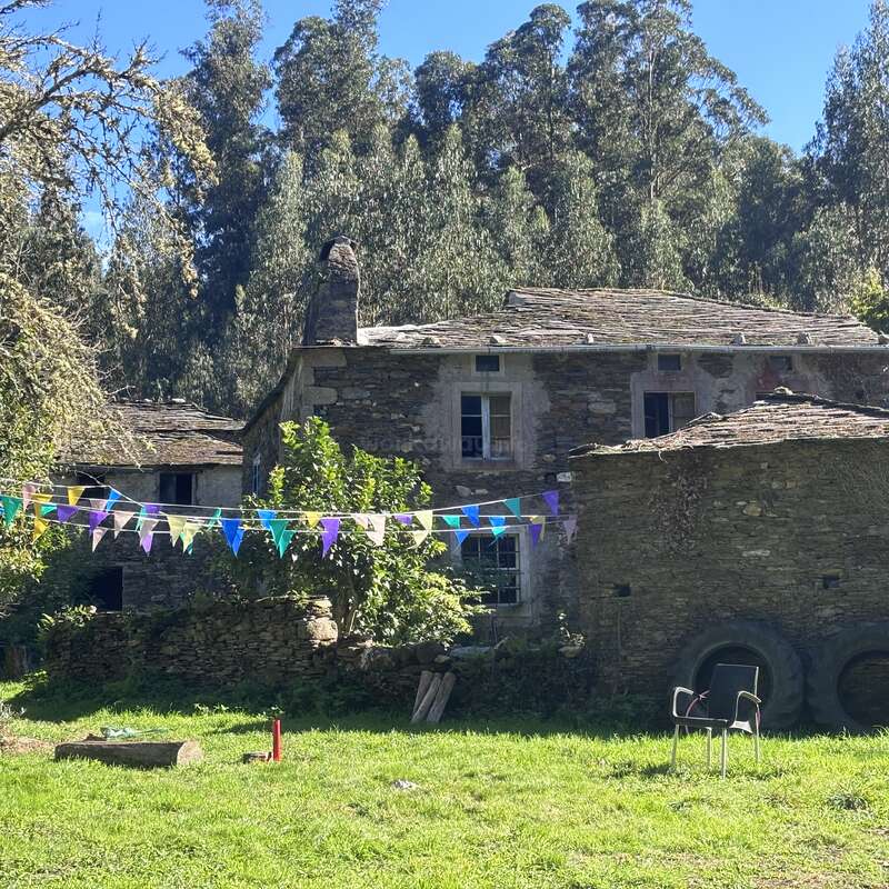 Uma antiga casa de pedra fica sob a luz do sol, cercada por uma vegetação exuberante. Uma faixa triangular colorida está pendurada sobre o quintal gramado, com uma cadeira e pneus por perto.