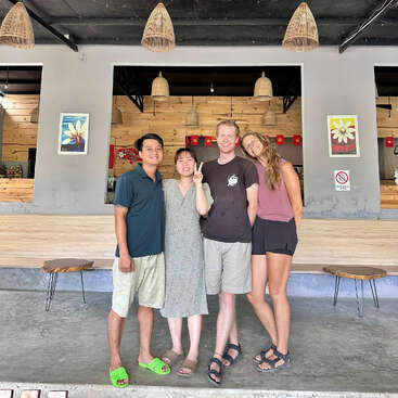 Cuatro amigos están muy juntos, sonriendo frente a una cafetería moderna con decoración de madera y lámparas colgantes. Dos cuadros con motivos florales decoran la pared de color gris claro.