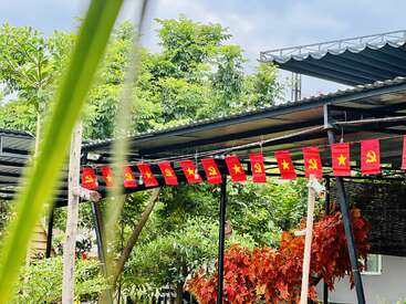 Rote Fahnen mit gelben Sternen und Hammer-und-Sichel-Symbolen hängen von einem Dach in einem üppigen Garten mit grünen Bäumen, bunten Blättern und einem Metallunterstand.