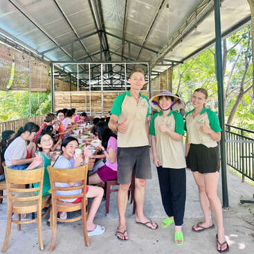 Um grupo de crianças está sentado à mesa comendo, enquanto três adultos, vestindo camisetas iguais, estão em pé na frente, sorrindo e fazendo sinal de "joinha" em uma área coberta ao ar livre.