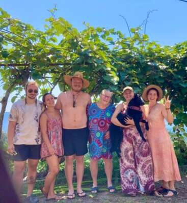 Six people and a black dog pose happily outdoors under green vines, enjoying sunny weather. They are casually dressed, smiling, and embracing each other, radiating warmth.