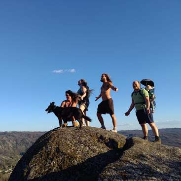 A group of four adults, one child in a backpack carrier, and a black dog stand joyfully atop a rocky hill under a clear blue sky.