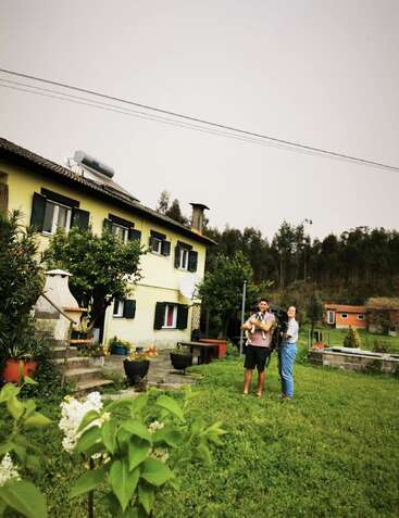 A couple stands smiling in a lush green garden outside a yellow house. One holds a dog, surrounded by plants, trees, and countryside with cozy vibes.