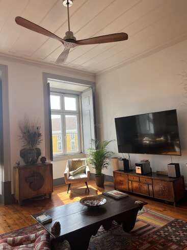 This cozy living room features a wooden ceiling fan, large window, TV, wooden furniture, potted plant, patterned rugs, and warm natural sunlight streaming in.