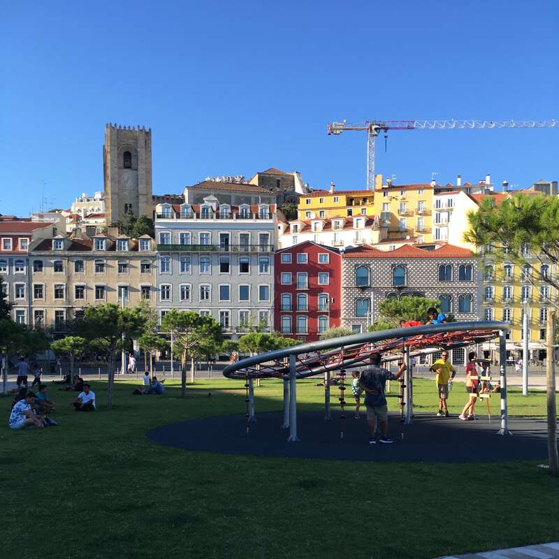 Children play on playground equipment in a park, surrounded by people relaxing, vibrant colorful buildings, a church tower, and a construction crane under a clear blue sky.