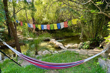 Una colorida hamaca cuelga entre los árboles junto a un tranquilo arroyo. Sobre el agua, rodeadas de un exuberante follaje verde y la luz del sol, cuelgan vibrantes banderas de oración.