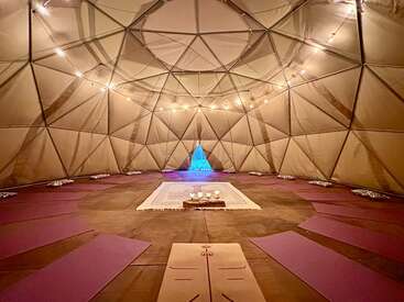 Un acogedor interior de cúpula geodésica con esterillas de yoga dispuestas en círculo, iluminación tenue, velas sobre una alfombra central y un altar azul resplandeciente al fondo.