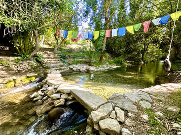Un sereno arroyo fluye bajo coloridas banderas de oración. Unos escalones de piedra y un pequeño puente invitan a explorar. La luz del sol se filtra entre la exuberante vegetación, creando un remanso de paz y naturaleza.