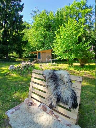 Un banc rustique en bois recouvert d'une peau de mouton trône sur l'herbe verte. Derrière, une petite cabane en bois est entourée d'arbres et d'une végétation luxuriante baignés de soleil.