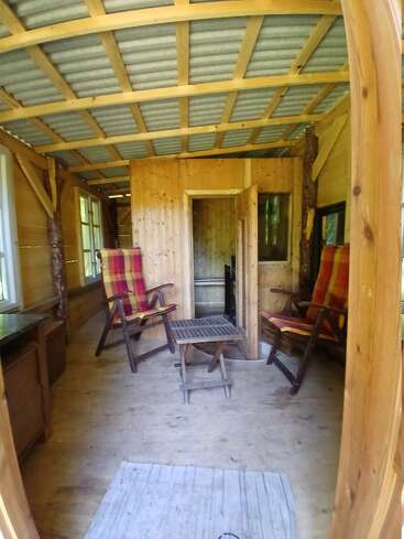 Cette image montre l'intérieur chaleureux d'une cabane en bois, avec deux fauteuils rembourrés à carreaux, une petite table, des murs en bois, des fenêtres ouvertes et un plafond rustique non fini.