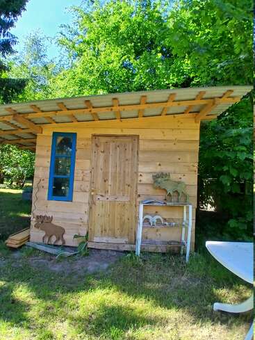 Une petite cabane en bois se dresse dans un jardin ensoleillé, entourée d'arbres verdoyants. Ornée de silhouettes d'animaux et d'une fenêtre bleue, elle dégage une atmosphère rustique et chaleureuse.