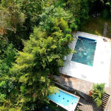 This aerial image shows a rectangular swimming pool and a smaller, narrower pool surrounded by dense green trees and foliage, with a stone path nearby.