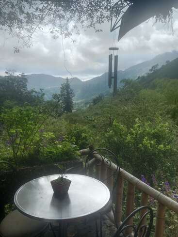 A serene patio with a metal table and chairs overlooks lush green hills and mountains, under cloudy skies. Hanging wind chimes add a peaceful touch.