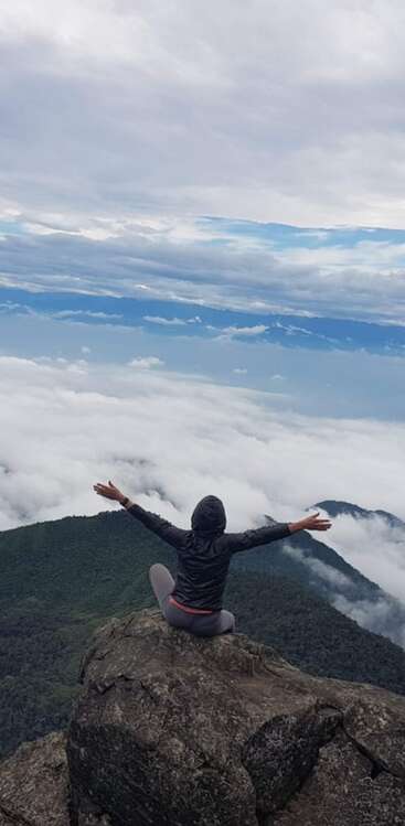 The image depicts a person sitting on a rocky outcrop, arms outstretched, overlooking a mountainous landscape with a cloudy sky. The person is dressed in a black jacket and grey pants.