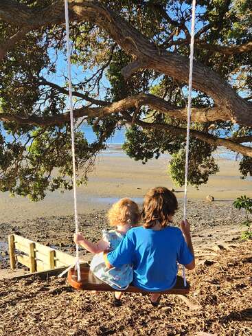 Duas crianças estão sentadas juntas num balanço pendurado numa árvore grande, com vista para uma praia tranquila. A luz do sol penetra por entre as folhas, criando uma cena tranquila e comovente.