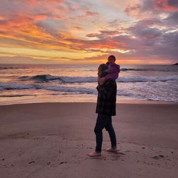 Um homem caminha descalço pela praia ao pôr do sol, carregando uma criança nos ombros. O céu brilha com tons vibrantes de laranja, rosa e roxo.