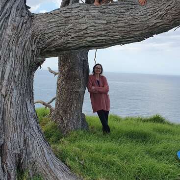 The image depicts a serene scene of a woman and two children on a tree branch overlooking the ocean, with a cloudy sky and lush green grass in the foreground.