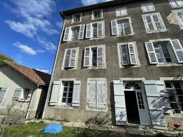 This image shows an old, three-story house with many white shuttered windows, aged walls, blue sky, some clouds, and a small, detached building nearby.