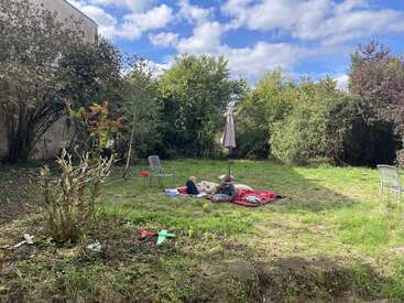 Two children lie on blankets in a sunny backyard, surrounded by green grass, trees, garden chairs, and toys. A peaceful, relaxing outdoor afternoon scene.