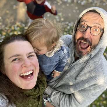 A joyful family selfie outdoors. A woman, man in glasses and hoodie, and two children. Smiles, laughter, and happiness captured in a candid, loving moment together.